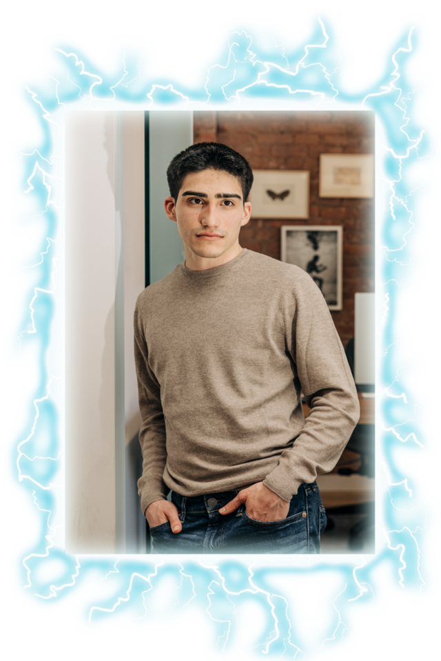 A young man with dark hair leans against a wall, wearing a beige sweater and blue jeans, with framed art in the background and a glowing blue electric border.